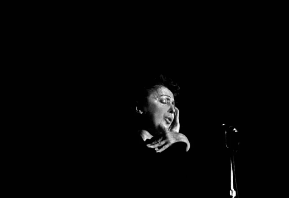 A imagem captura um instante de suspensão. Piaf canta quase sem se mover, como se toda a ação estivesse concentrada na voz. O fundo negro não é ausência: é silêncio. A mão junto ao rosto sugere esforço, entrega, intimidade. Não há espetáculo — há presença. É o retrato de uma artista que transformou fragilidade em força expressiva e deu forma sonora a um tempo ferido.